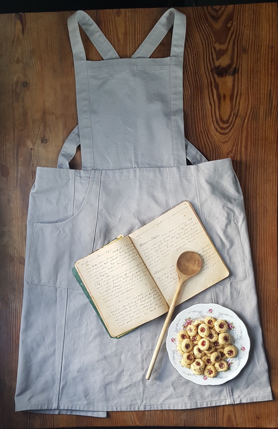 graue Latzschürze vor Holzhintergrund. Altes Rezeptbuch, Holzkochlöffel und ein Teller mit Plätzchen.
