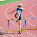Eine Hürdenläuferin mit wehenden blonden Haaren und Sonnenbrille springt über eine Hürde.