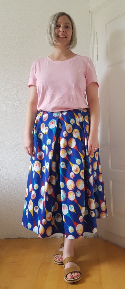 Galaoutfit Sabine in einem rosafarbenen T-Shirt und einem blau-gemusterten, wadenlangen Rock, der in der Taille in unregelmäßige Falten gelegt ist.