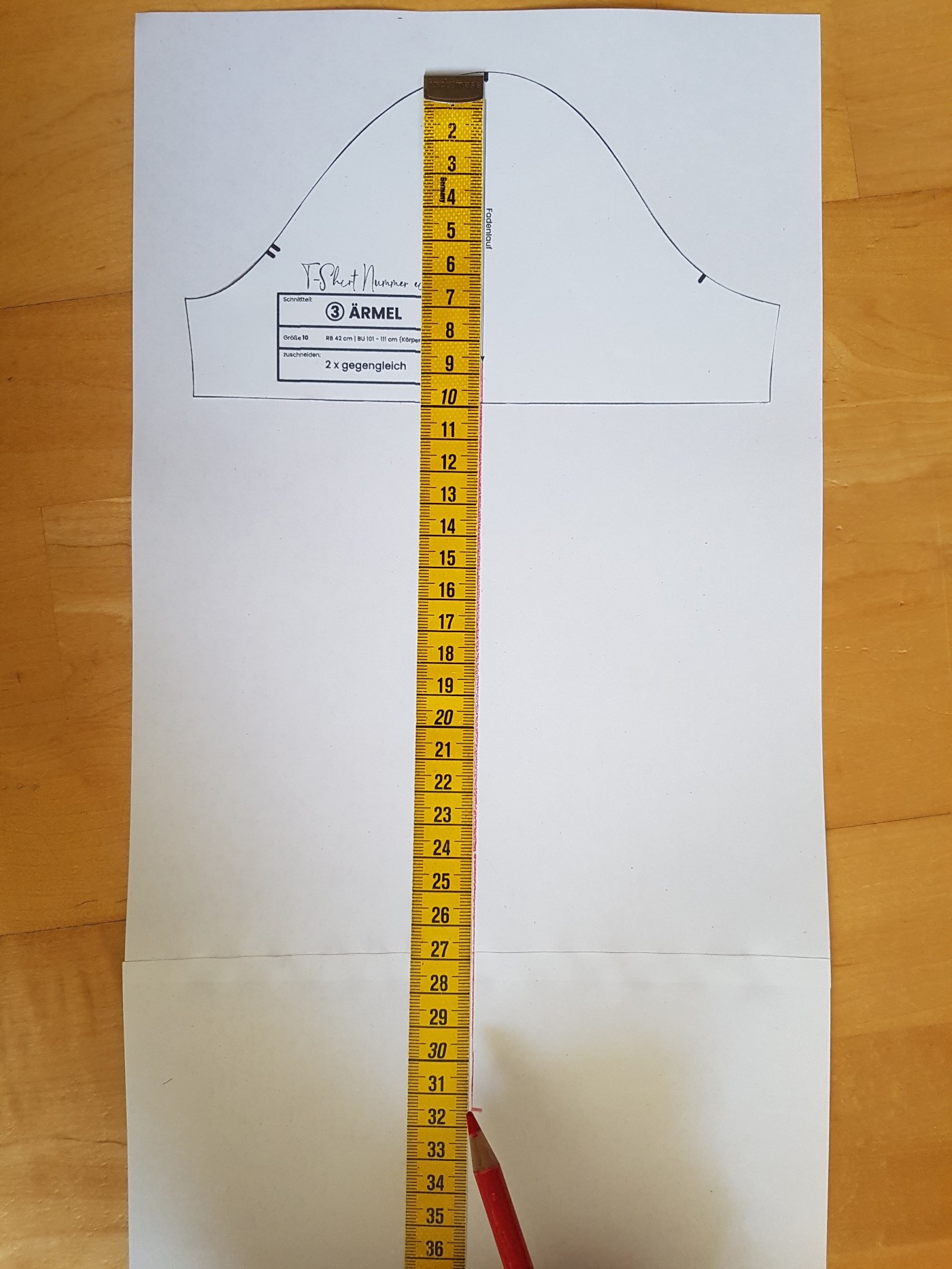 Der Ärmelschnitt liegt auf einem größeren Papier. Ein Maßband zeigt an, wo die Mittellinie verlängert und die Ärmellänge abgetragen wird. Das abgebildete Schnittmuster hat einen Maßstab von 2:1