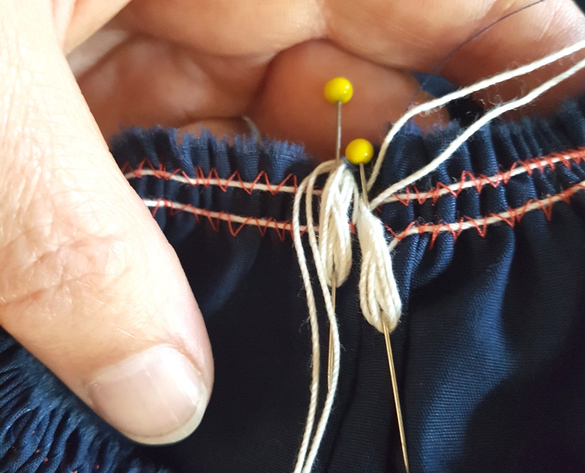 Eine Hand hält den angereihten Stoff. Die beiden Reihfäden sind in 8er-Schlaufen um Stecknadeln gewickelt, um das Reihgarn zu fixieren.
