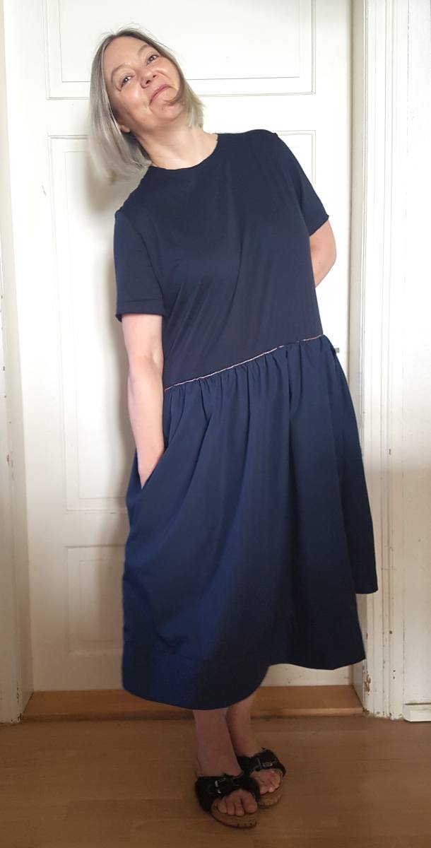 Sabine steht im neuen Kleid vor einer weißen Tür und muss sich tief zur Seite beugen, um die Hand in den Taschenbeutel zu stecken. Immerhin - sie lacht!