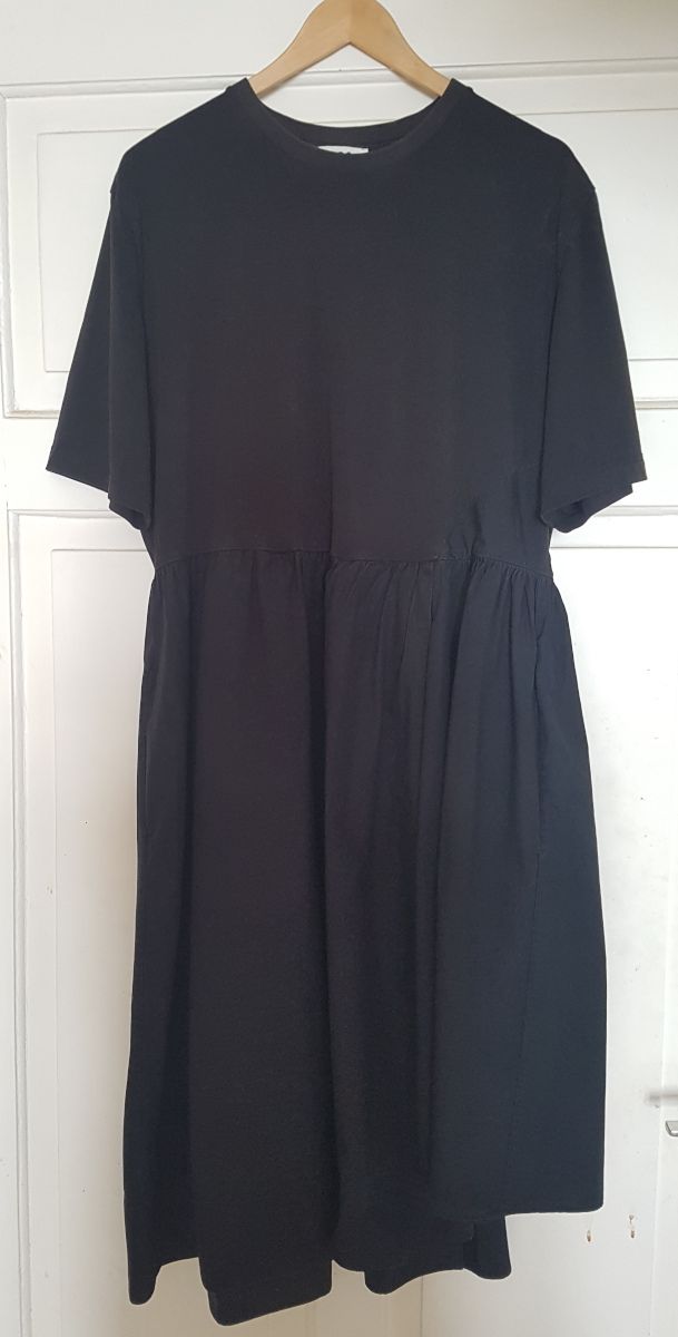 Ein schwarzes Kleid hängt auf dem Bügel an einer Tür. Das Oberteil sieht aus wie ein normales T-Shirt mit Rundhalsausschnitt, der Rockteil ist weit, angekräuselt  und aus Webstoff.
