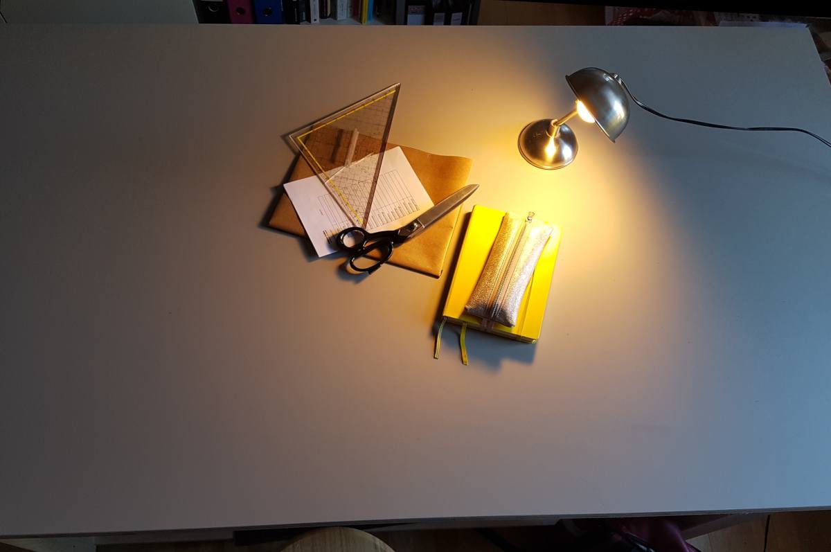 Die Tischplatte von oben. Auf dem Tisch steht eine kleine Schreibtischlampe, ein gelbes Notizbuch, eine Zuschneideschere, ein Geodreieck und ein paar Zettel.