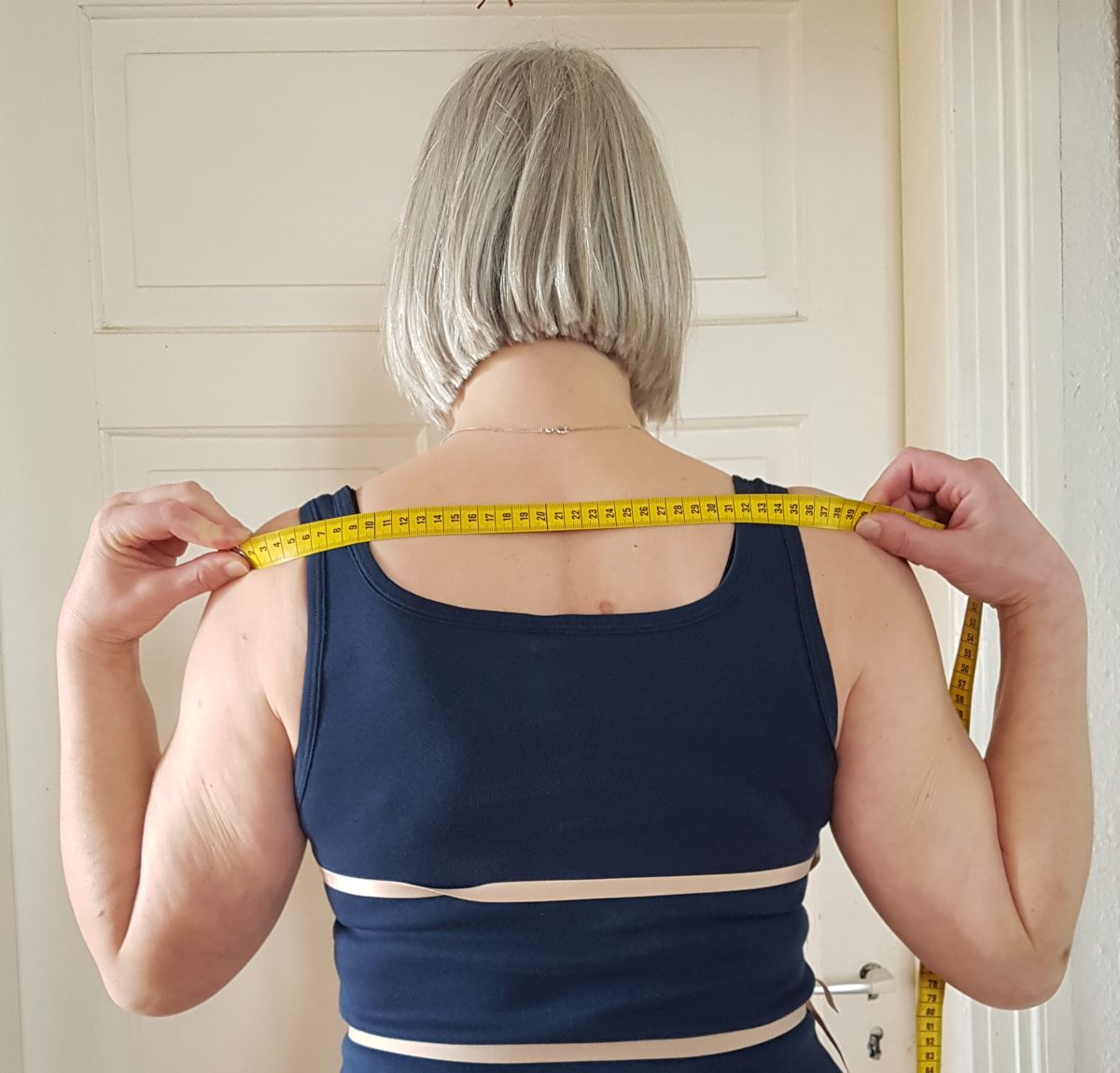 Schulterbreite messen Sabine von hinten. Das Maßband liegt quer über dem oberen Rücken, von Schulterknochen zu Schulterknochen
