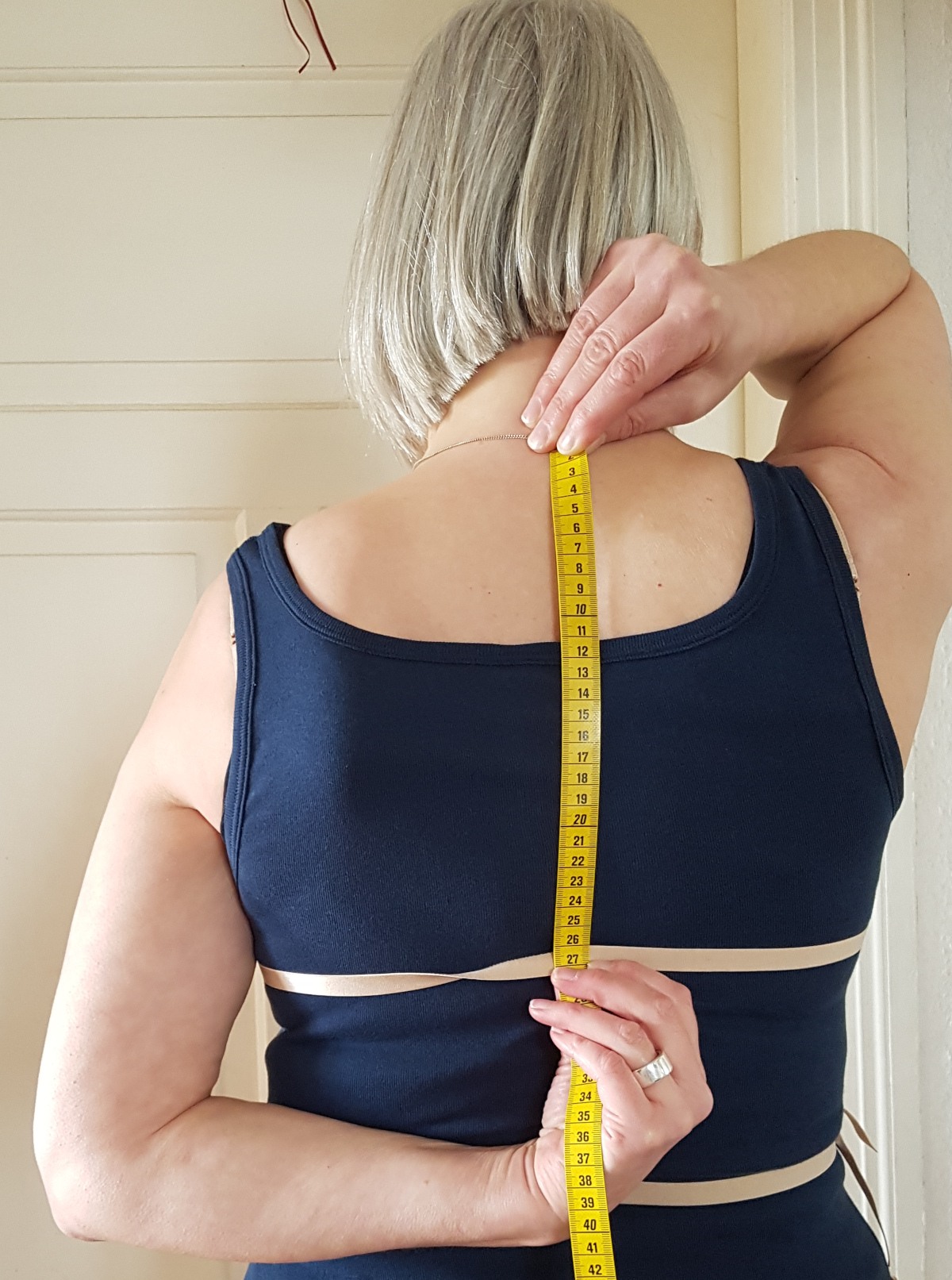 Rückenhöhe messen Sabine von hinten. Sie hält das Maßband mit der rechten Hand an den Halsansatz und markiert mit der linken Hand die Höhe der Brustlinie