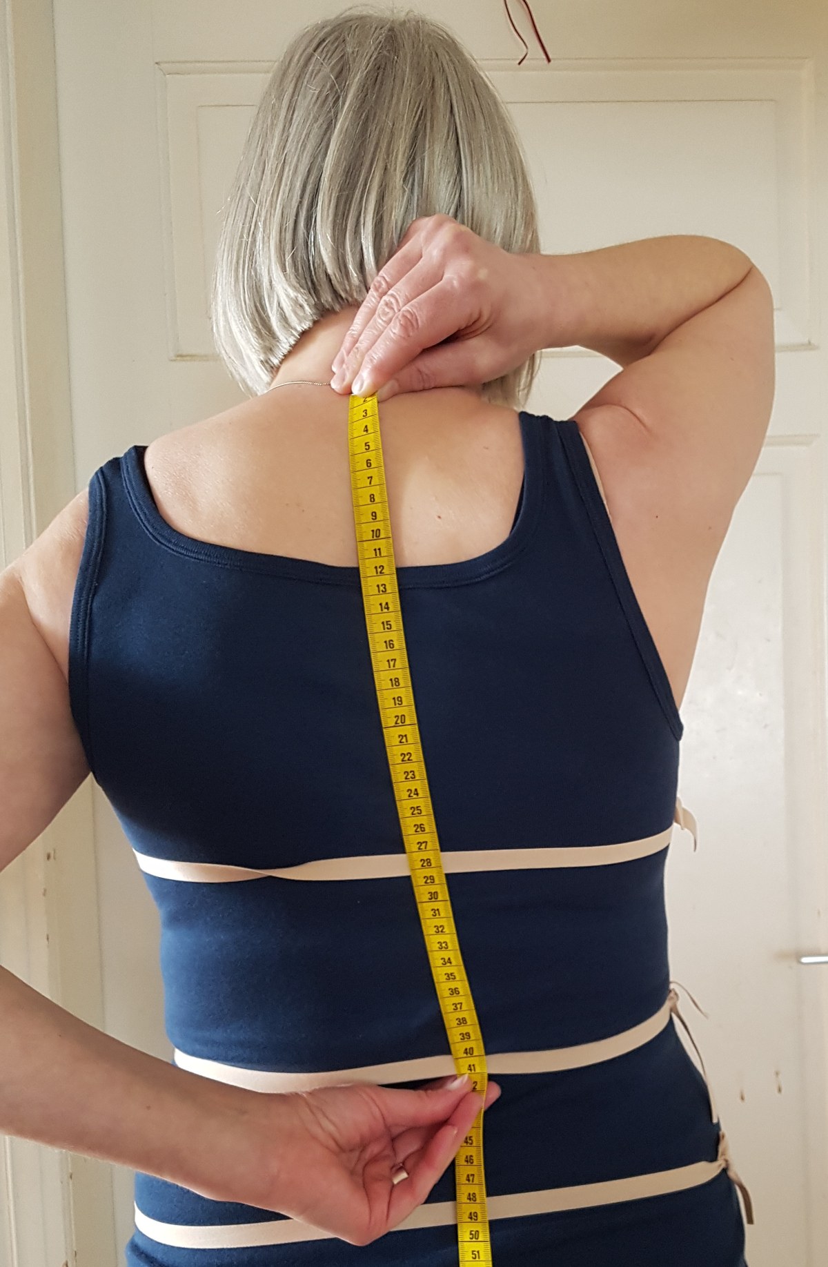 Rückenlänge messen Sabine von hinten. Sie hält das Maßband mit der rechten Hand an den Halswirbel und mit der linken Hand an die Taille.