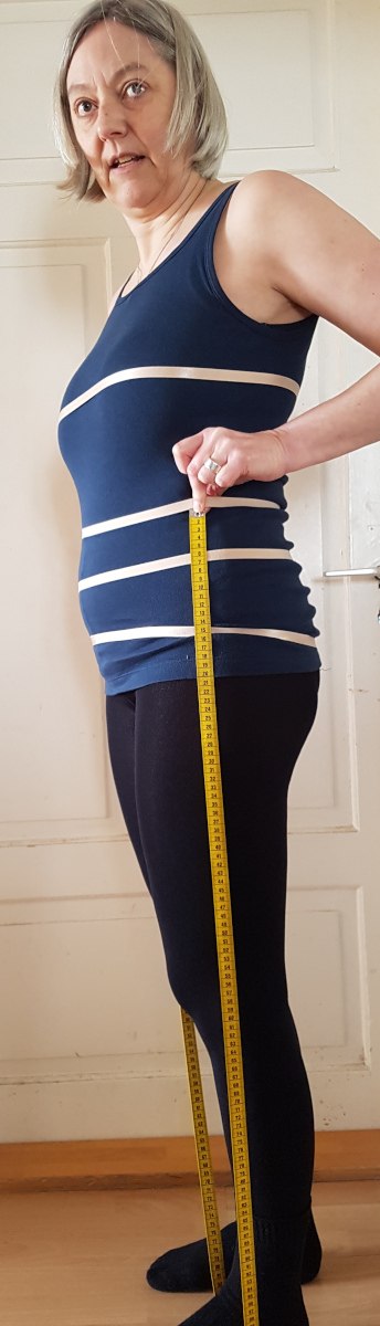 Seitenlänge messen Sabine in Seitenansicht. Sie hält den Anfang des Maßbands auf Taillenhöhe und steht mit dem Fuß auf dem Maßband am Boden. Das Maßband ist gespannt.